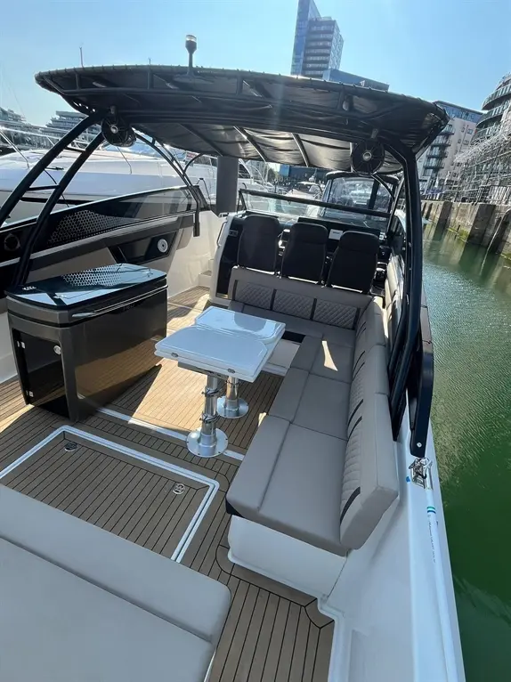 Luxury boat interior featuring grey cushioned seating, a central table, and a stylish black cooler, with a sunny marina backdrop.
